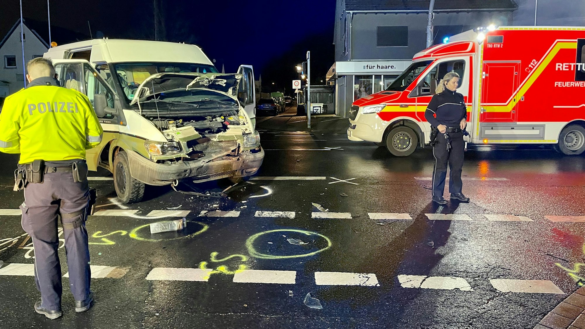 Auf der regennassen Fahrbahn, die mit aufgesprühten Markierungen versehen ist, steht ein Kleinbus, dessen Front zusammengedrückt ist. Zwei Polizisten und ein Rettungswagen stehen daneben.