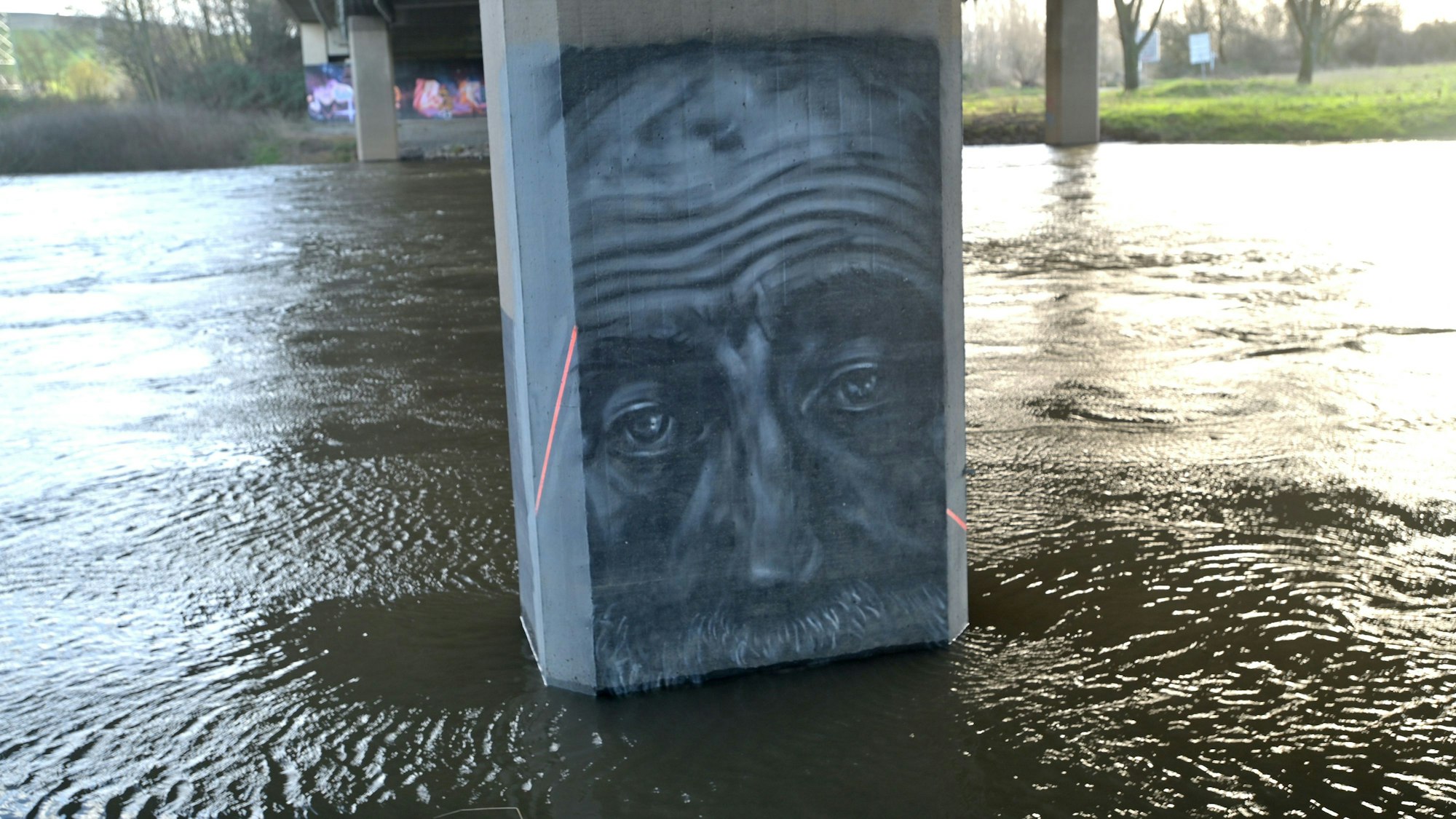 Wasser der Wupper umspült einen Brückenpfeiler, auf dem ein Graffiti einen Mann mit Schnurrbart zeigt.