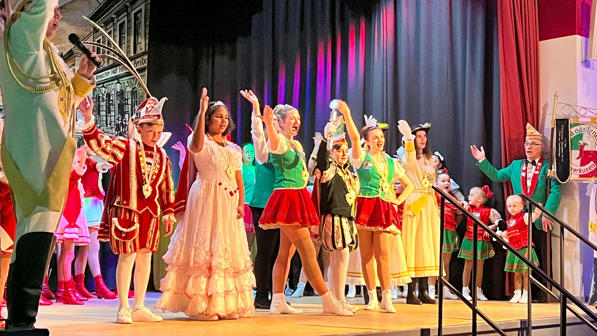 Ein Kinderprinzenpaar und ein Kinderprinz stehen in vollem Ornat auf der Bühne und interagieren mit dem Publikum. Um sie herum stehen andere Karnevalisten.