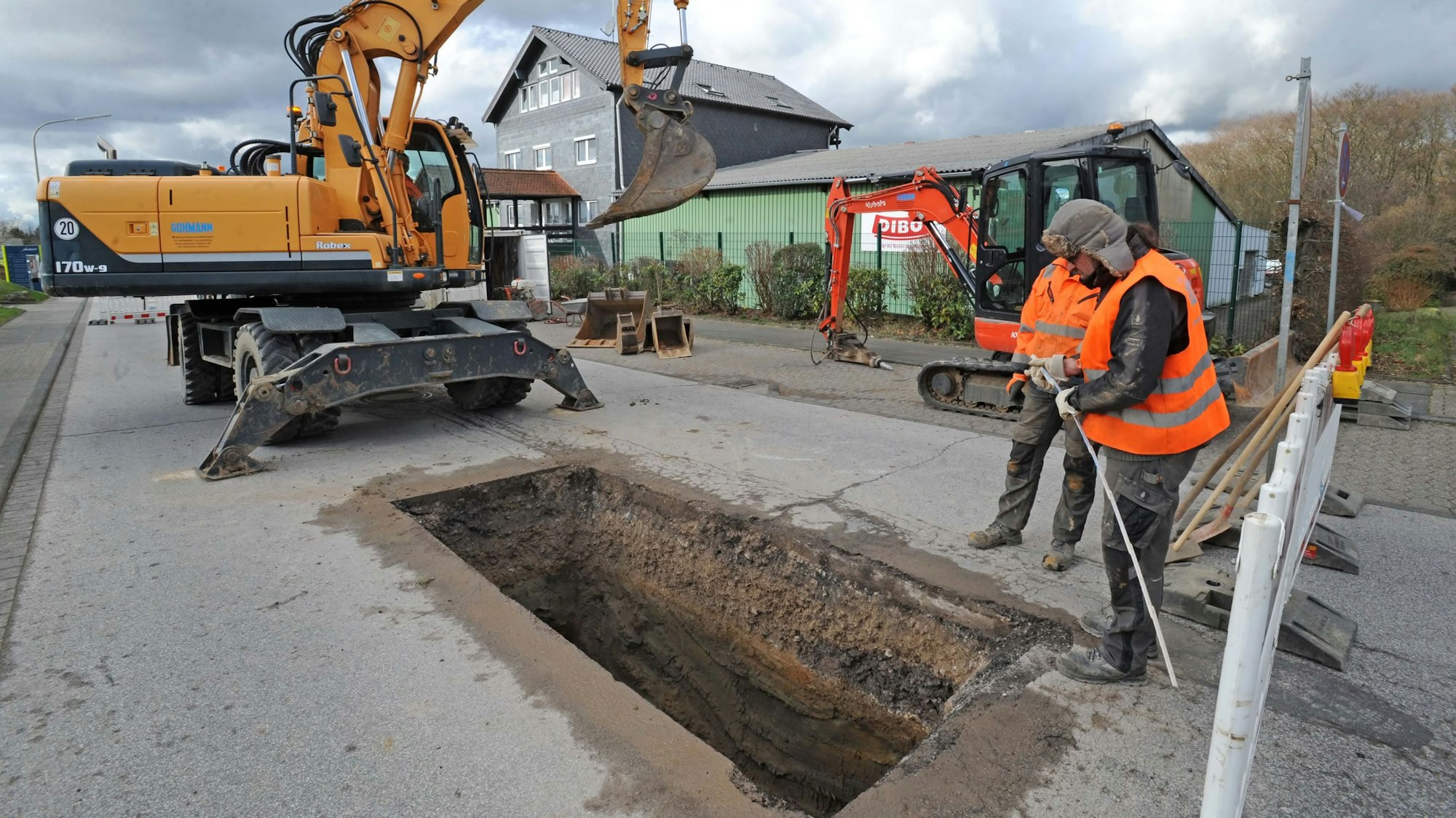Zwei Bauarbeiter stehen neben einem Bagger an einer Kanalbaustelle, die Straße ist aufgerissen.