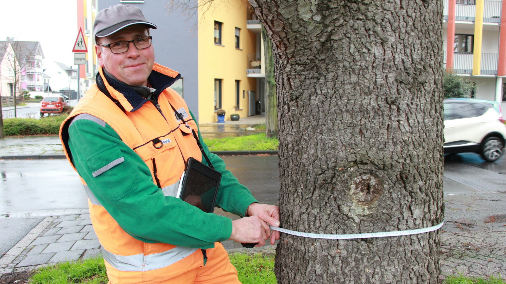 Ein Mann mit oranger Jacke hält ein Maßband um einen Baumstamm.