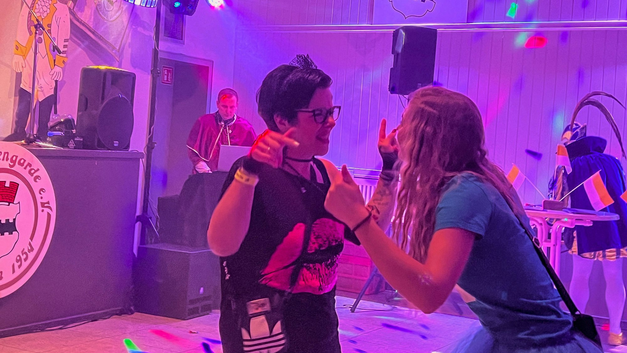 Zwei junge Frauen tanzen und singen, im Hintergrund ist ein DJ zu sehen.