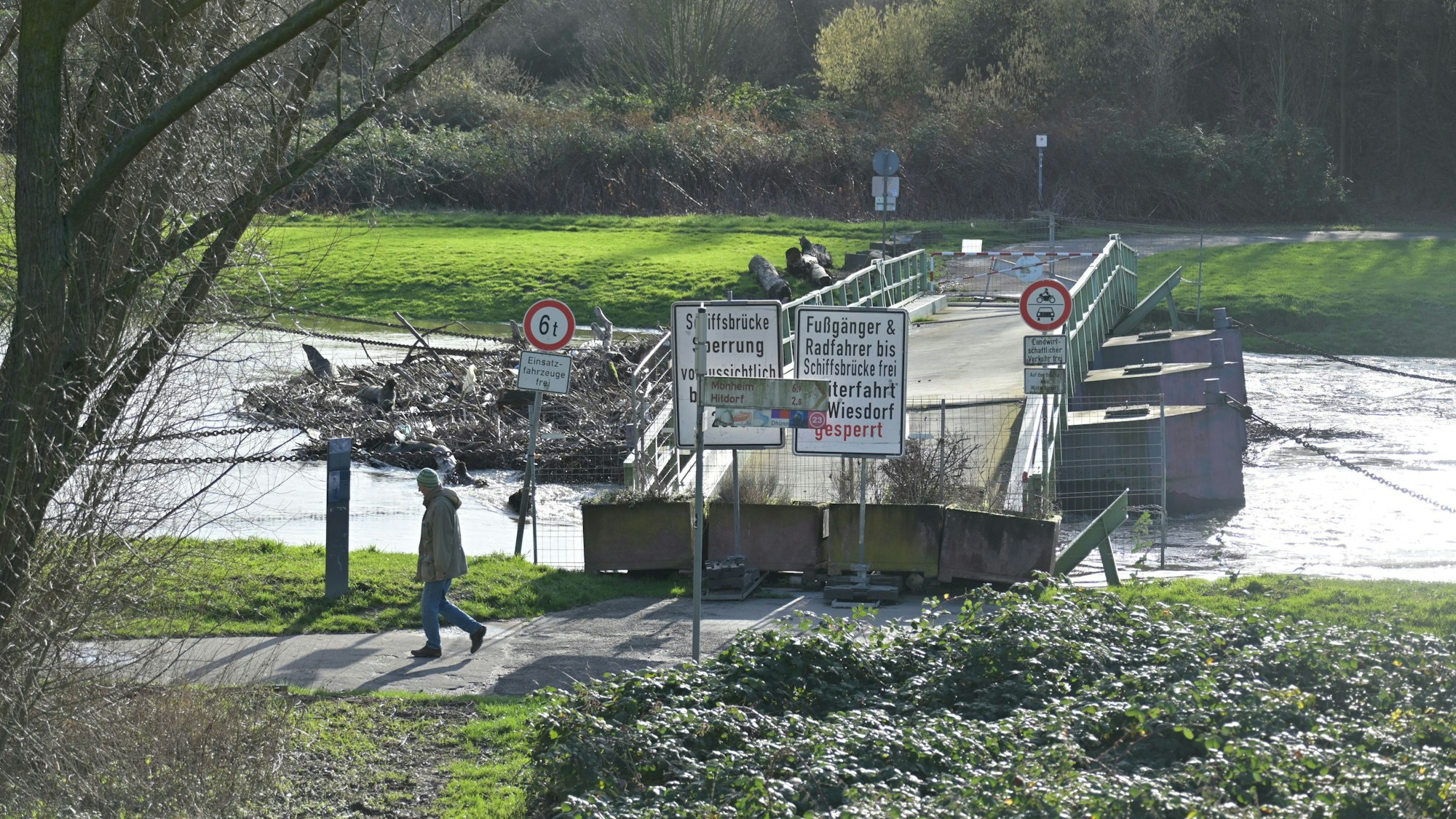 An der Pontonbrücke spaziert ein Mann vorbei. Die Brücke ist mit Bauzäunen abgesperrt. An einer Seite der Brücke sammeln sich Massen an Treibgut im Wasser.