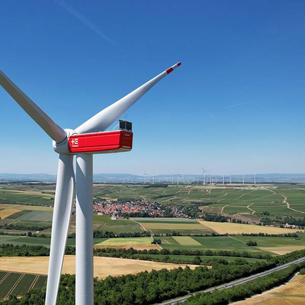 Vor einem weiten Horizont, Feldern und einer Ortschaft steht ein großes weiß-rotes Windrad.