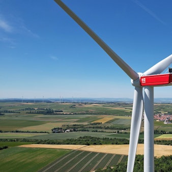 Vor einem weiten Horizont, Feldern und einer Ortschaft steht ein großes weiß-rotes Windrad.