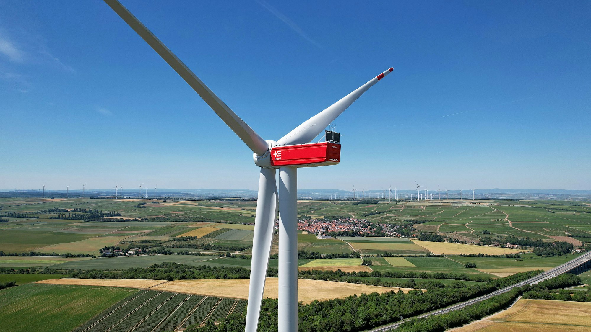 Vor einem weiten Horizont, Feldern und einer Ortschaft steht ein großes weiß-rotes Windrad.