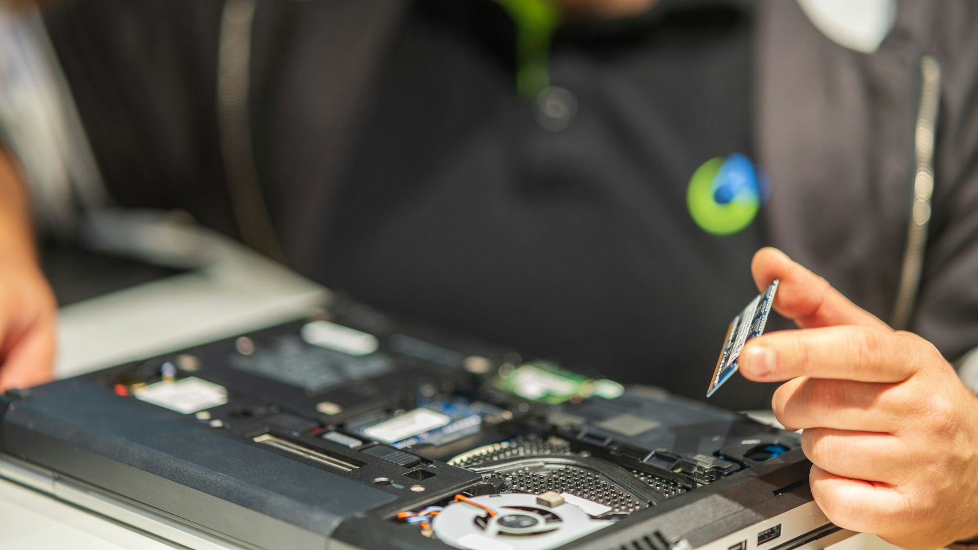 Ein aufgeschraubter Laptop liegt auf einem Tisch. Eine Person hält eine zugehörige Festplatte.