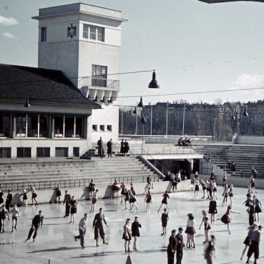 Menschen auf der Eisfläche. Links hinten im Bild ein Turm mit Hakenkreuz darauf.