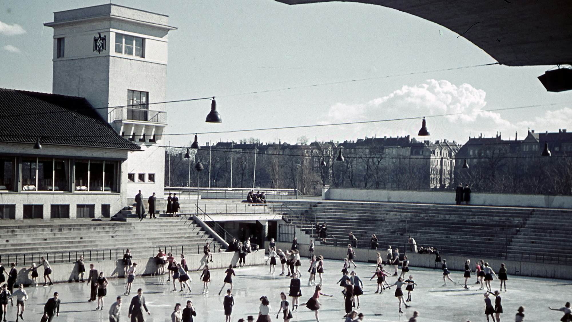 Menschen auf der Eisfläche. Links hinten im Bild ein Turm mit Hakenkreuz darauf.