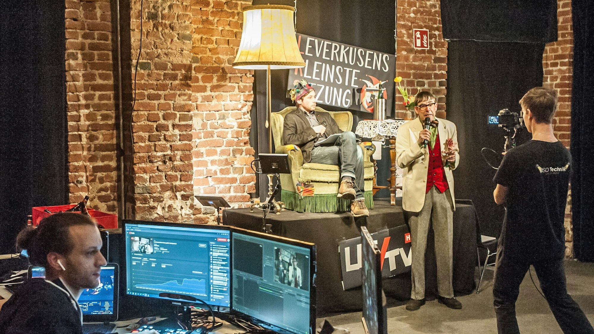 Leute vom Team der LKS sitzen an Tischen vor Monitoren oder stehen kostümiert bei Leverkusens Kleinster Sitzung im Kulturausbesserungswerk.