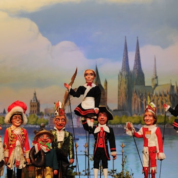 Stockpuppen des Hänneschentheaters vor dem Kölner Rheinpanorama: Rote Funken und Hellige Knäächte un Mägde posieren mit Speimanes und Sitzungsleiter Schäng.