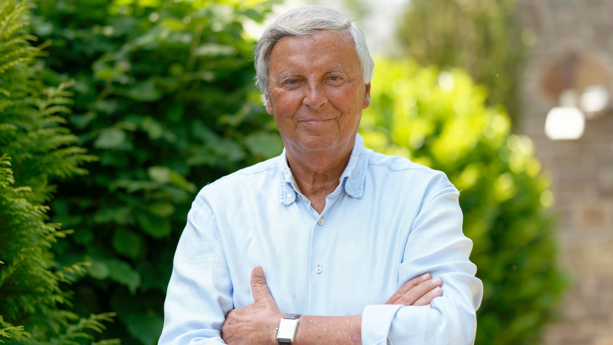 Wolfgang Bosbach hat die Arme vor der Brust überkreuzt und steht vor Büschen.