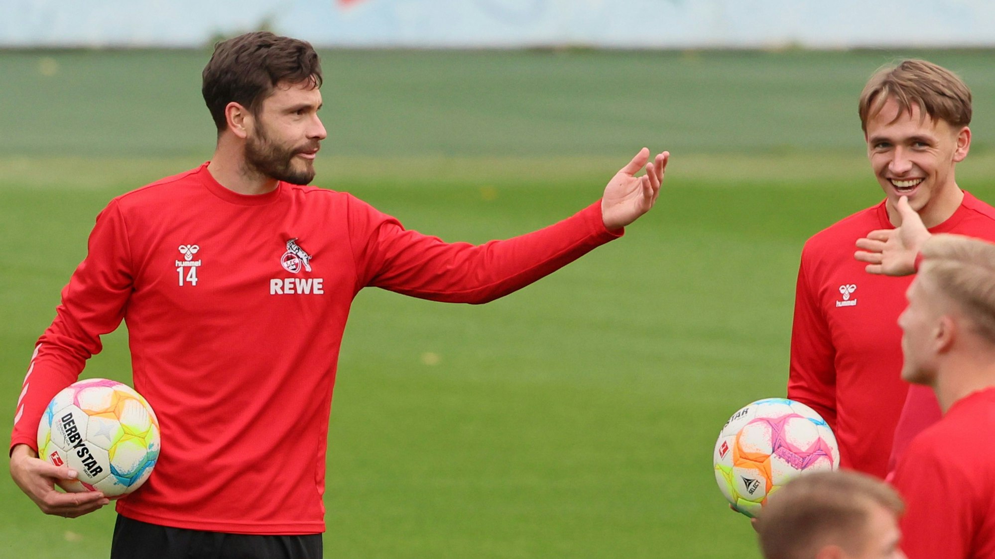 FC-Kapitän Jonas Hector gestikuliert während des Trainings mit dem Ball unter dem Arm. Kollege Mathias Olesen sieht ihm lachend zu.