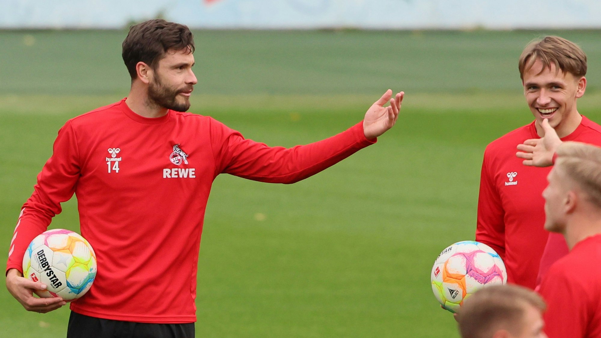 FC-Kapitän Jonas Hector gestikuliert während des Trainings mit dem Ball unter dem Arm. Kollege Mathias Olesen sieht ihm lachend zu.