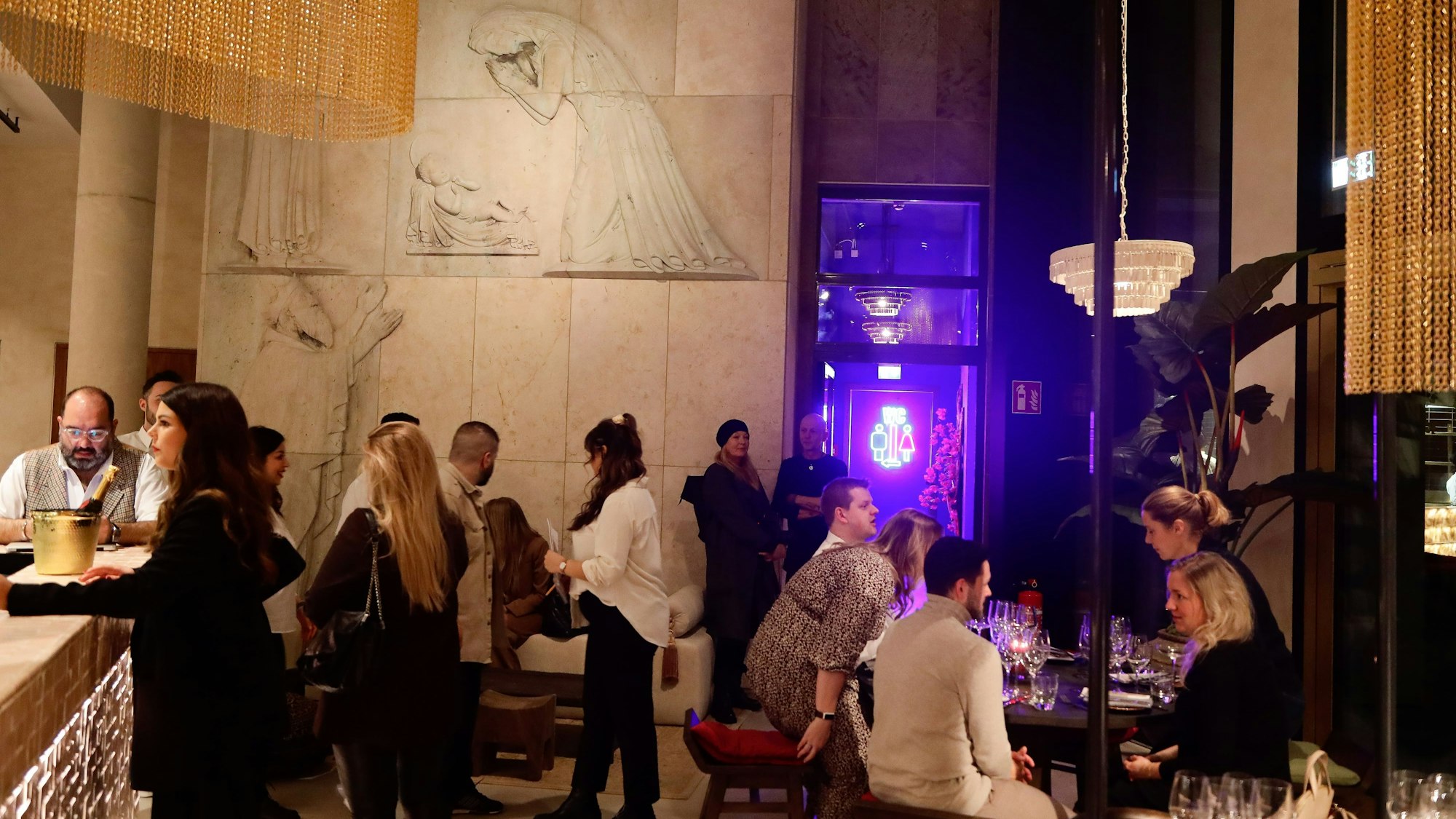 Gäste stehen im Raum. An der Wand hinter ihnen sind Figuren aus der Wand heraus gearbeitet.