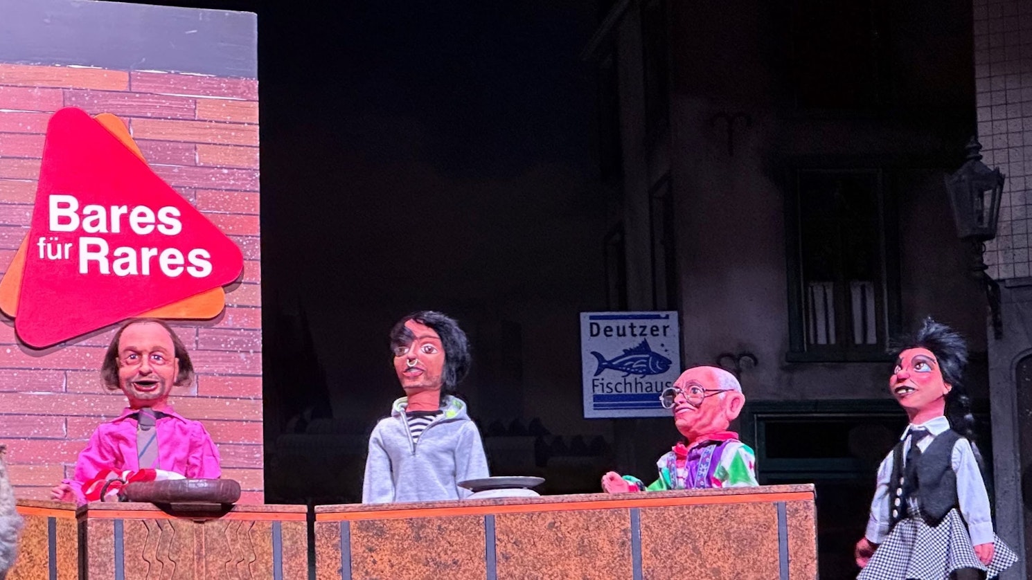 Röschen (r.) versucht bei „Bares für Rares“ für die Woosch einen guten Preis zu erzielen, links hinter Tischen die Experten der TV-Sendung, alle als Puppen.