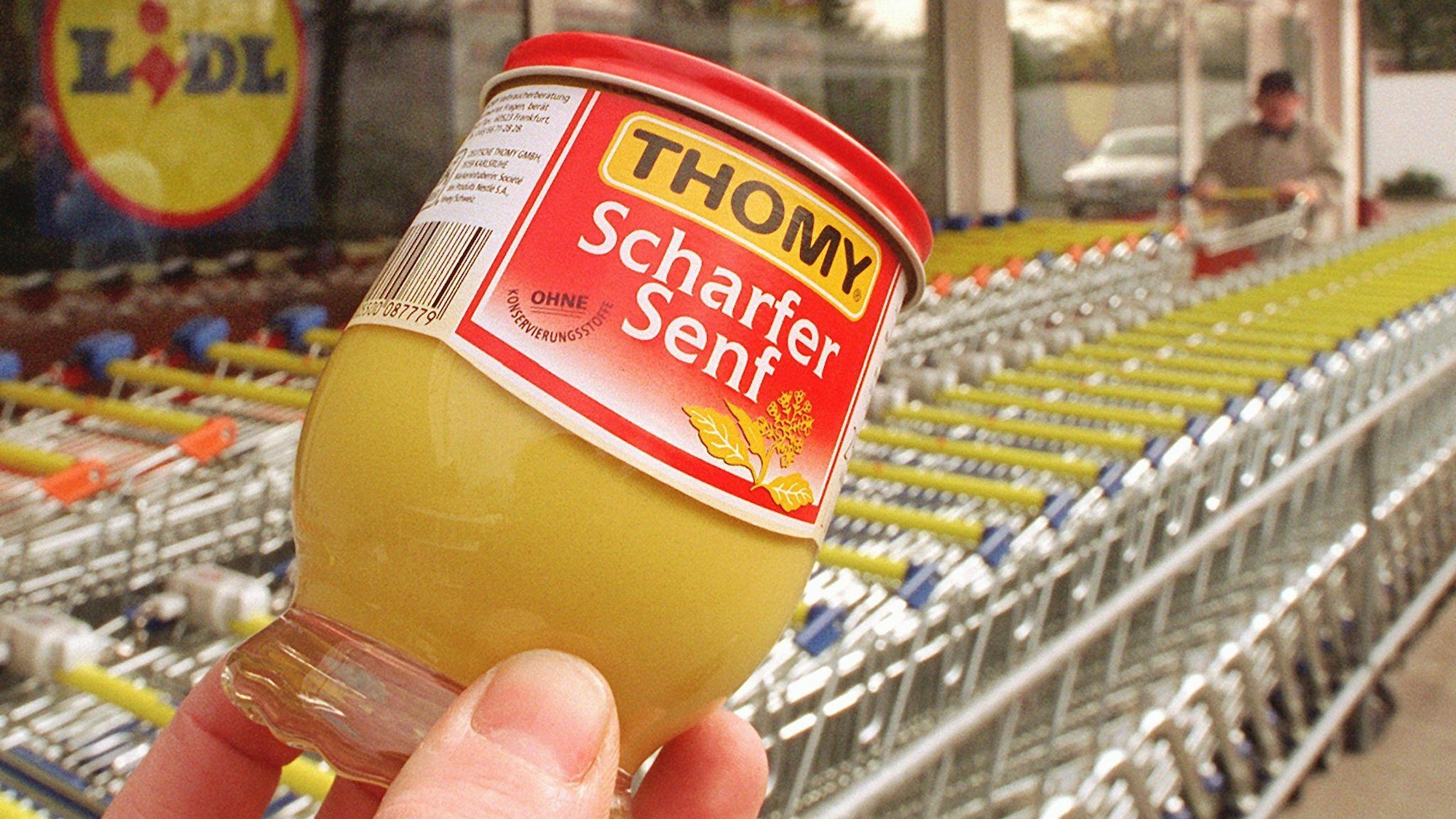 Vor abgestellten Einkaufswagen deines  Lidl-Lebensmittelgeschäftes wird symbolisch ein Senfglas der Firma Thomy ins Bild gehalten.