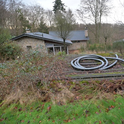Ein Haus steht auf einem verwucherten Grundstück, eine Bretterbude erhebt sich im Vordergrund.