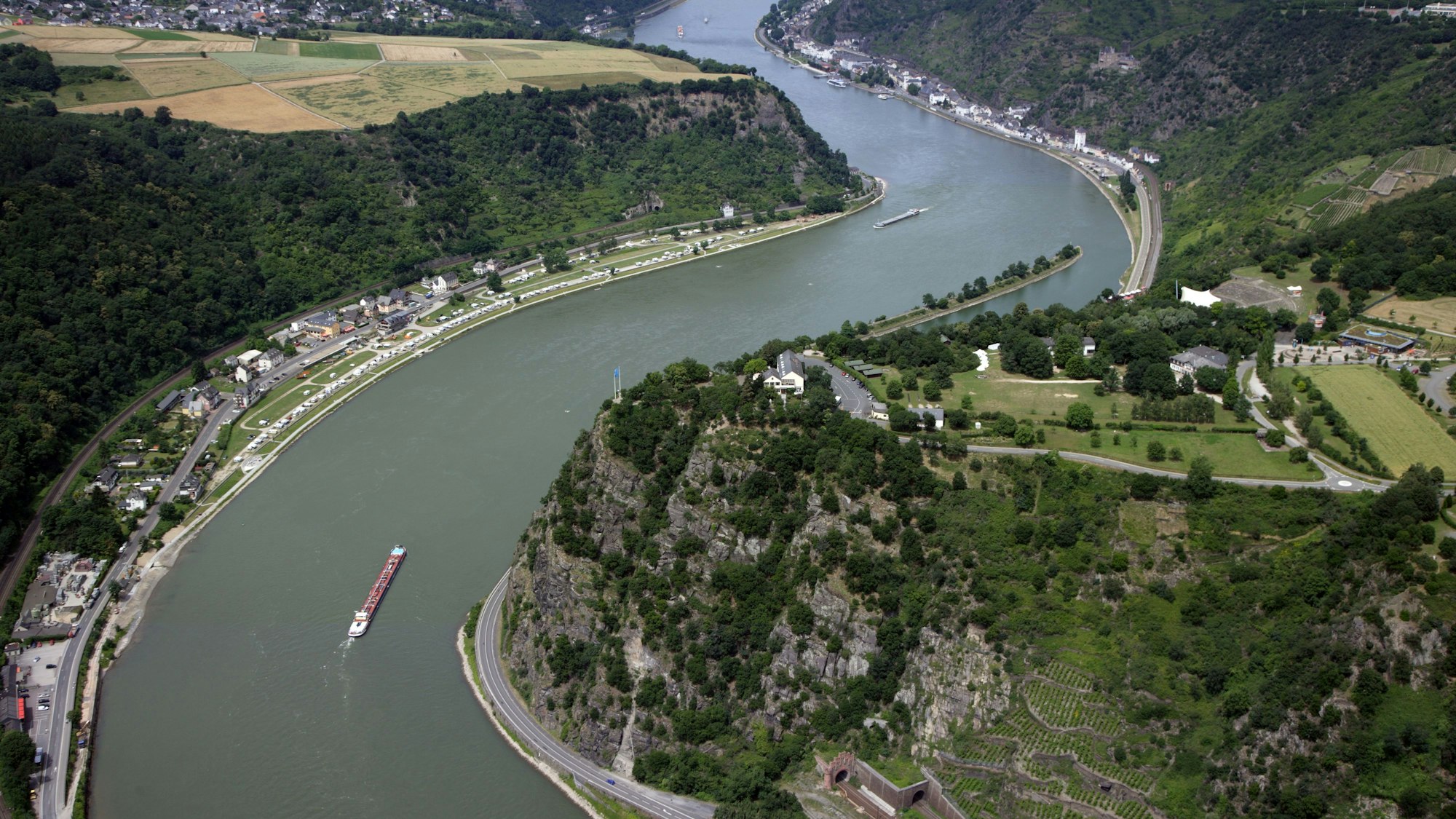 Der Rhein schlängelt sich durch die grüne Landschaft des Mittelrheintals beim Loreley-Felsen. Frachschiffte fahren auf dem Fluss.