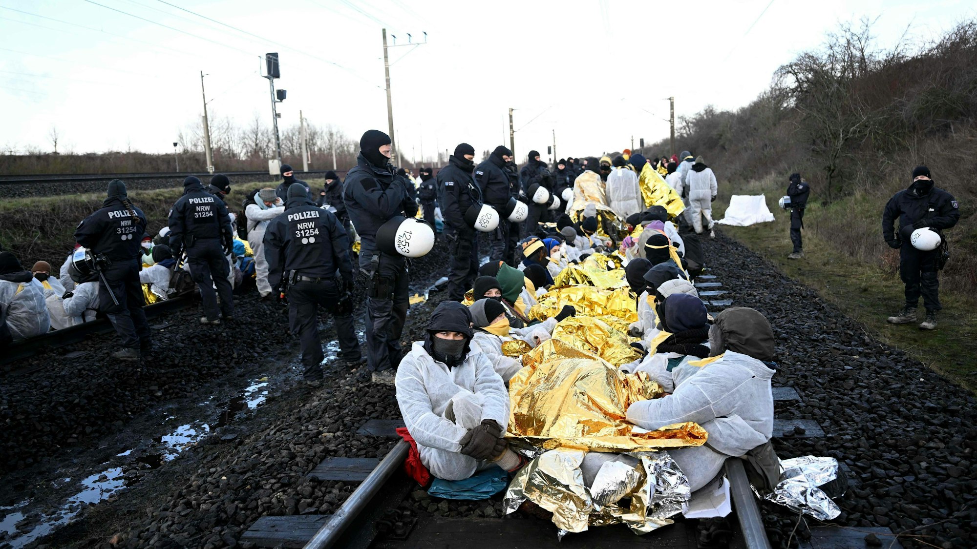Aktivisten in weißen Maleranzügen und Isolierfolie gehüllt sitzen auf Gleisen. Darum herum sind Einsatzkräfte der Polizei zu sehen.