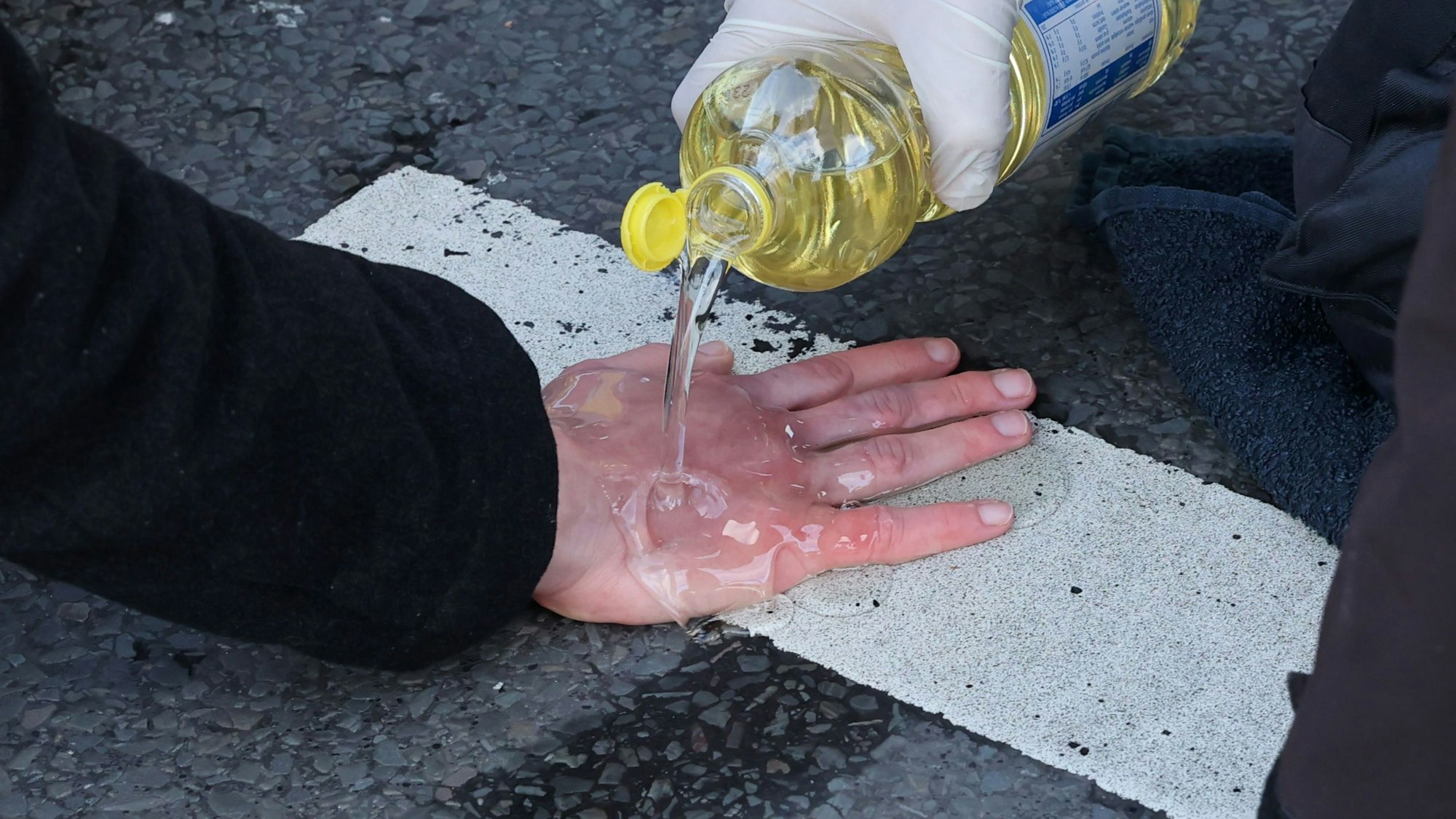 Eine an einer Fahrbahnmarkierung festgeklebte Hand wird von einem Rettungsdienstmitarbeiter mit Speiseöl übergossen.