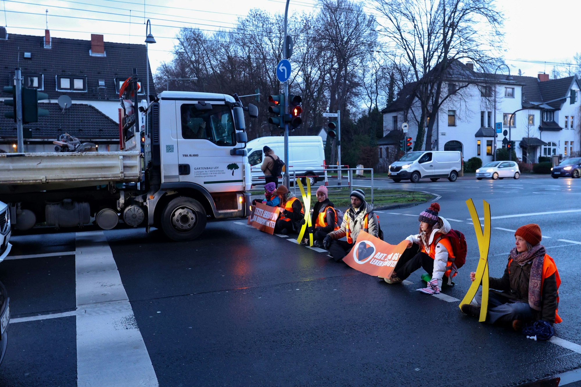 Klimaaktivisten sitzen auf der Aachener Straße an einer Ampel. Vor ihnen steht ein Lastwagen.
