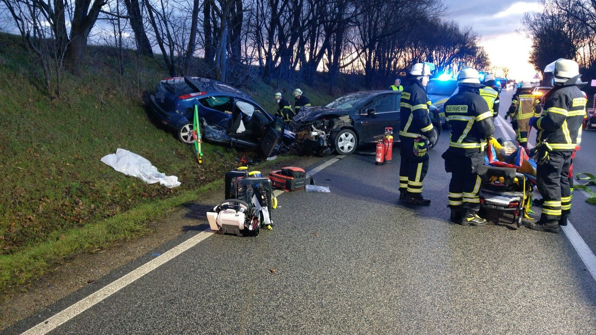 Mehrere Feuerwehrleute stehen an der Unfallstelle. Zwei Autos sind ineinander gefahren und stehen im Straßengraben.