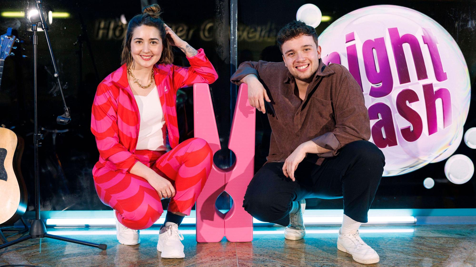 Luisa Charlotte Schulz und Ben Schafmeister hocken neben einer großen Wäscheklammer auf der Bühne vom Waschsalon, in dem Nightwash aufgezeichnet wird.