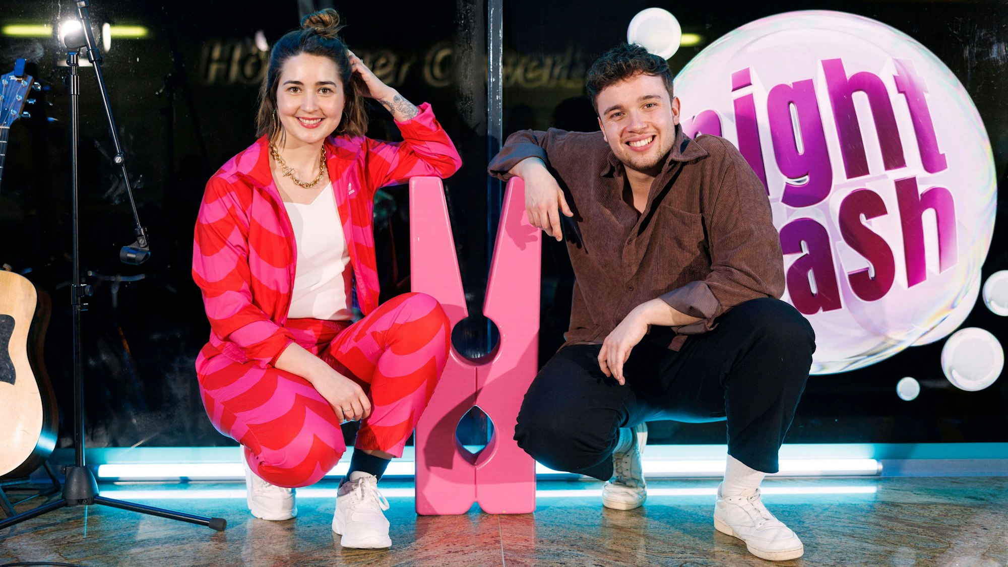 Luisa Charlotte Schulz und Ben Schafmeister hocken neben einer großen Wäscheklammer auf der Bühne vom Waschsalon, in dem Nightwash aufgezeichnet wird.