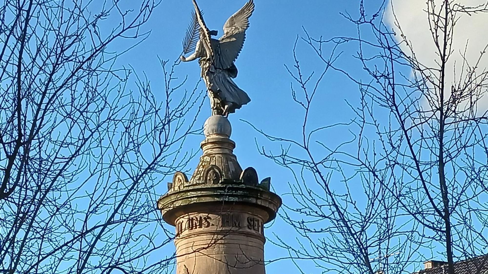 Eine steinerne Frauenfigur mit Flügeln und einer Feder in der Hand steht auf der Spitze einer Steinsäule.