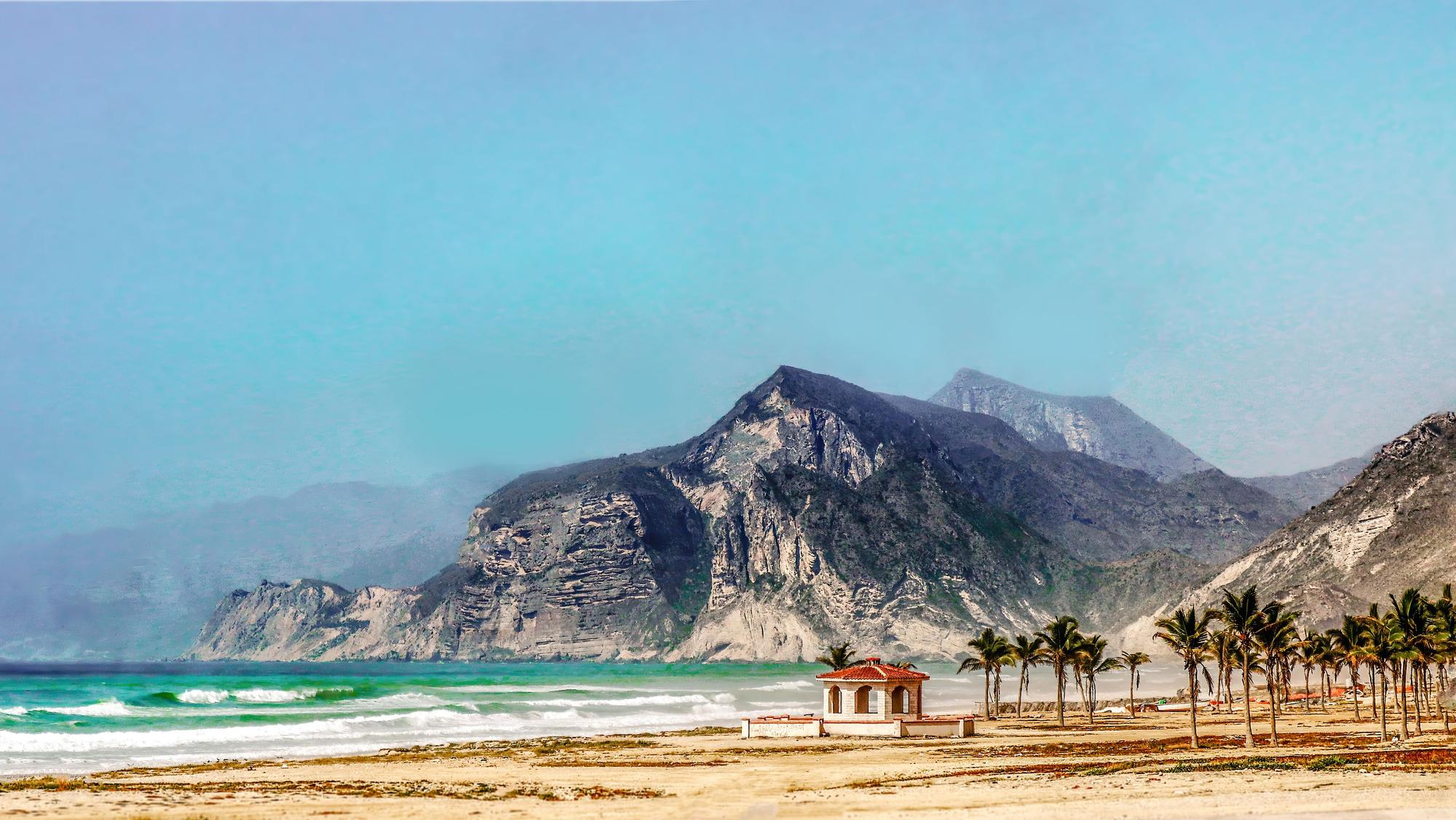 Am Strand von Slalah stehen Palmen und ein kleines Häuschen, im Hintergnd Berge