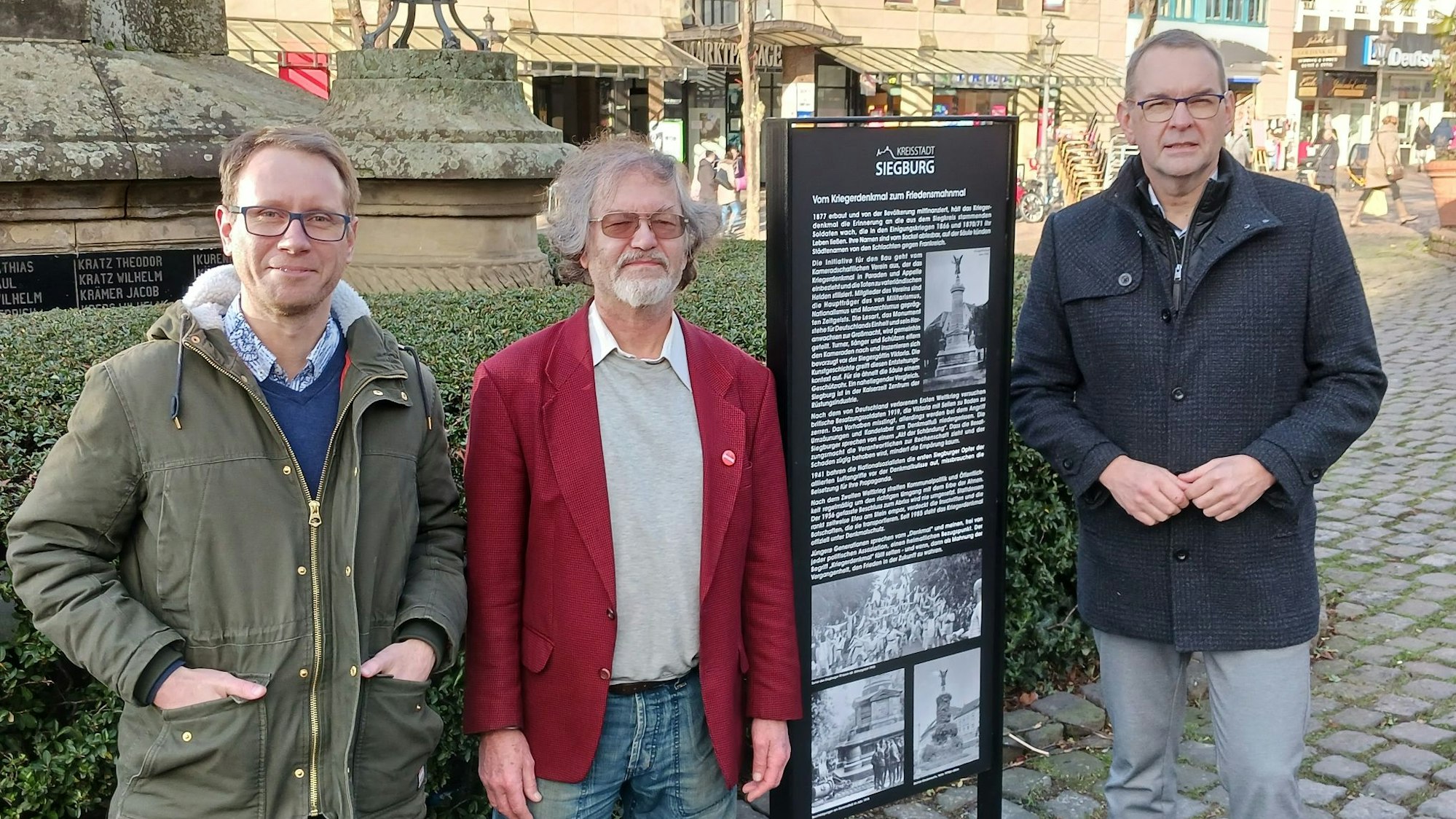 Drei Männer stehen neben einer Informationstafel, auf der Texte und alte Fotos abgedruckt sind.