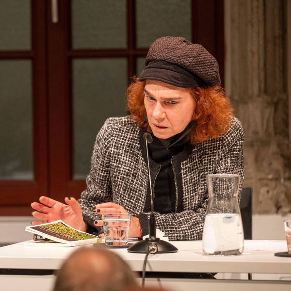 Aslı Erdoğan hat ihr Buch vor sich auf einem Tisch liegen liegen und spricht in ein Mikrofon. Sie hat rote Haare und trägt eine Mütze.