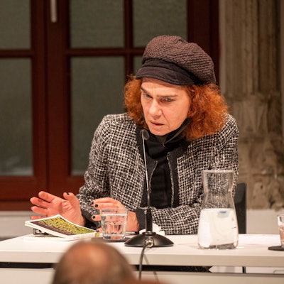 Aslı Erdoğan hat ihr Buch vor sich auf einem Tisch liegen liegen und spricht in ein Mikrofon. Sie hat rote Haare und trägt eine Mütze.