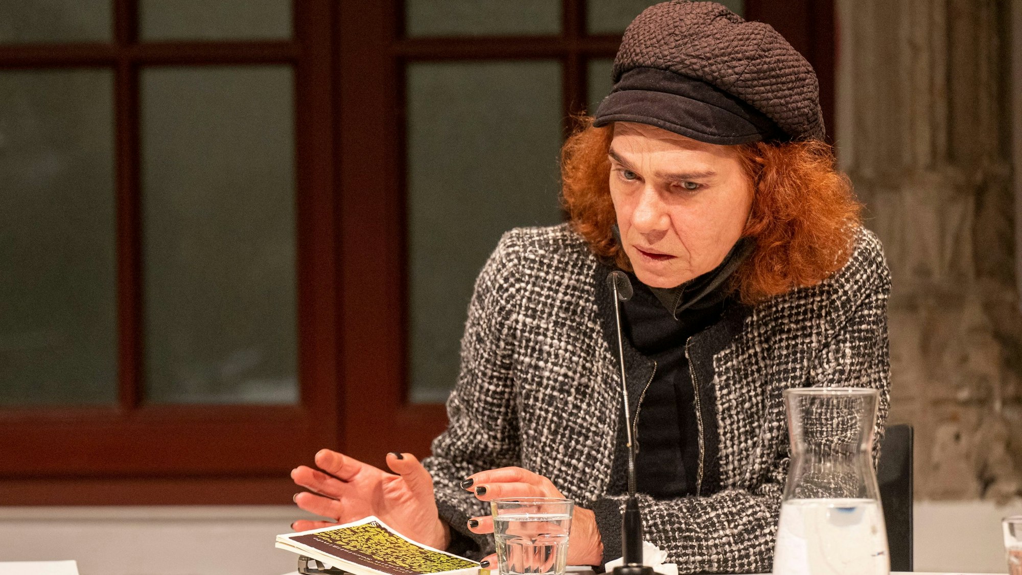 Aslı Erdoğan hat ihr Buch vor sich auf einem Tisch liegen liegen und spricht in ein Mikrofon. Sie hat rote Haare und trägt eine Mütze.