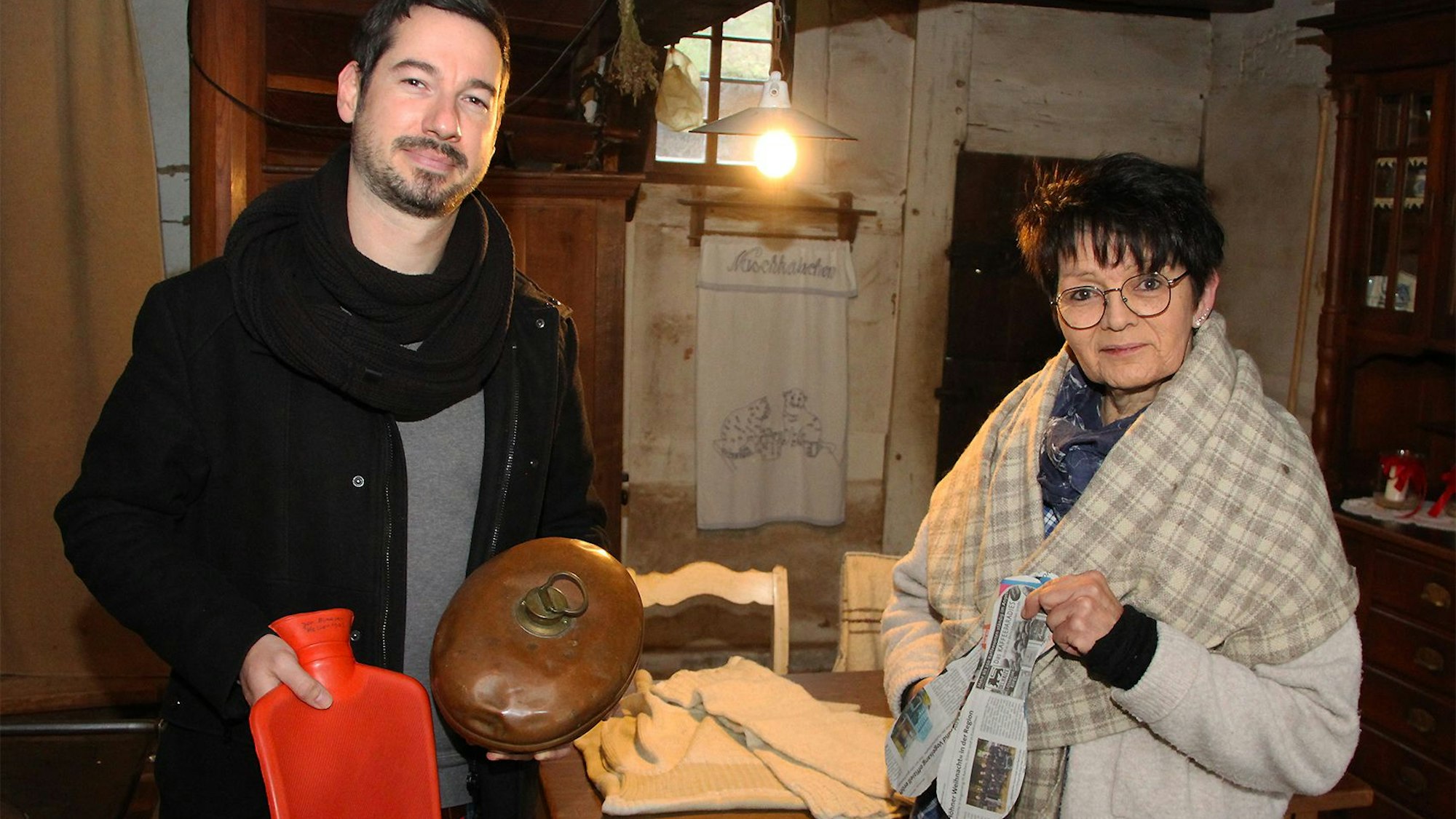 Ein Mann und eine Frau zeigen Hausmittelchen gegen die Kälte: Wärmflaschen, kupferne Bettwärmer und in Sohlenform geschnittenes Zeitungspapier.