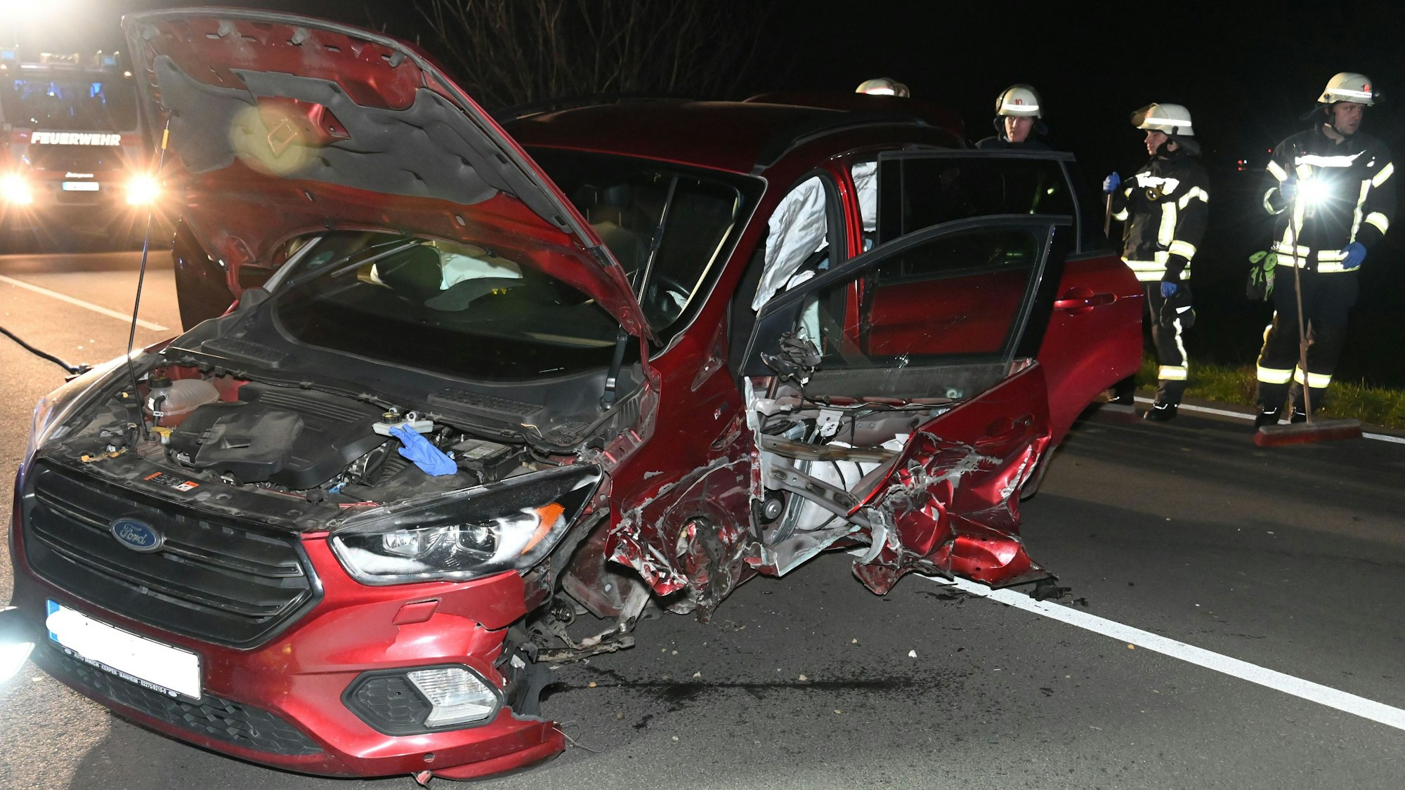 Ein rotes Auto ist nach einem Unfall komplett zerstört. Die Motorhaube steht offen. Im Hintergrund stehen Feuerwehrleute.