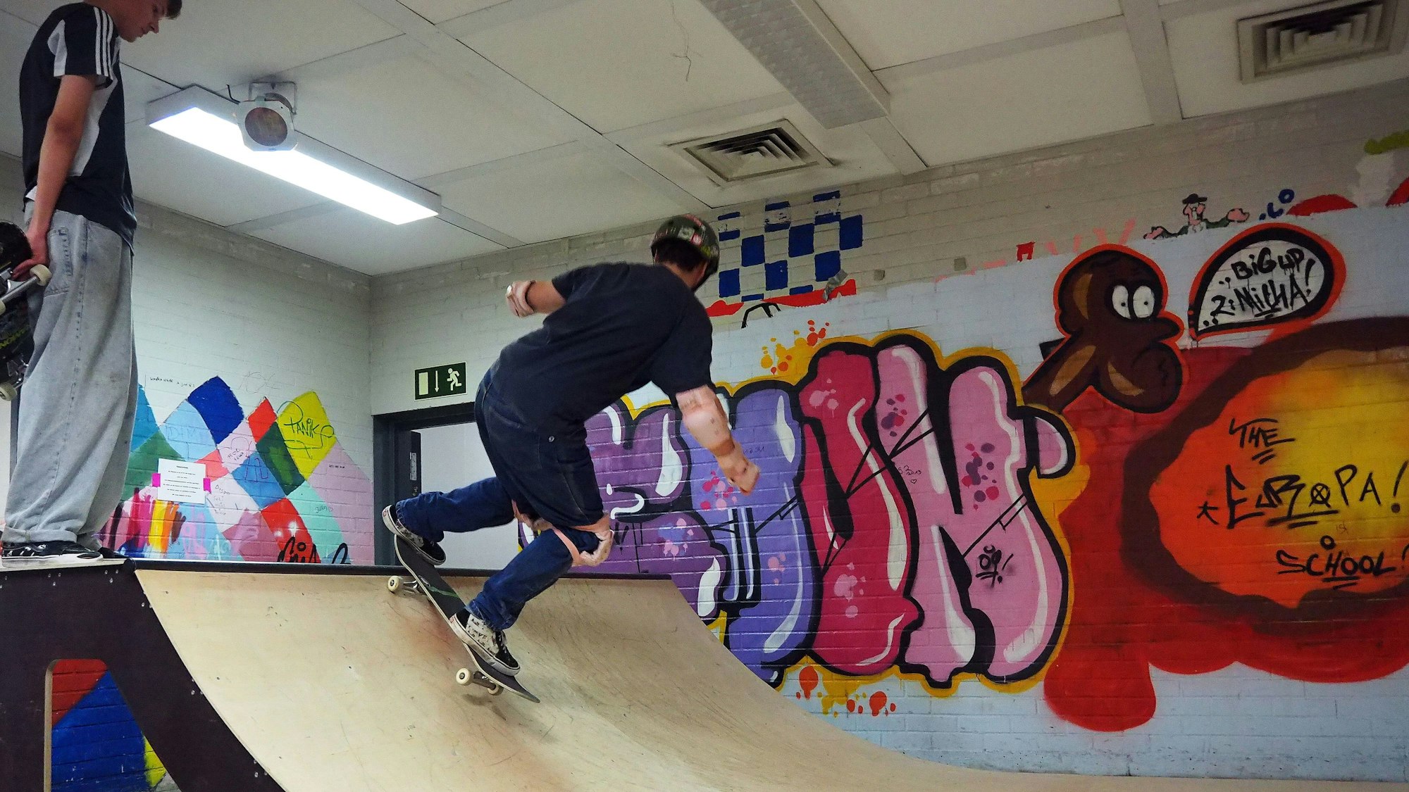 Schüler, der mit Helm und Schonern auf der Skateboard-Rampe fährt, während ein weiterer Schüler zuguckt