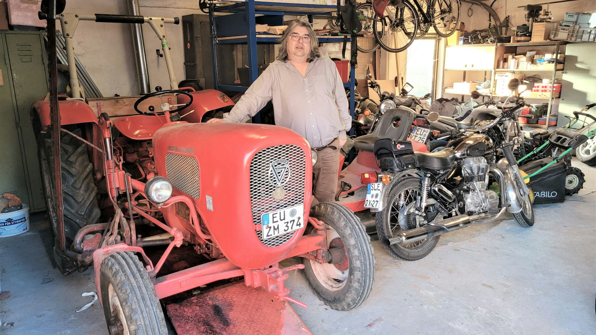 Ein Mann steht an einem alten Traktor, im Hintergrund sind einige Motorräder zu sehen.