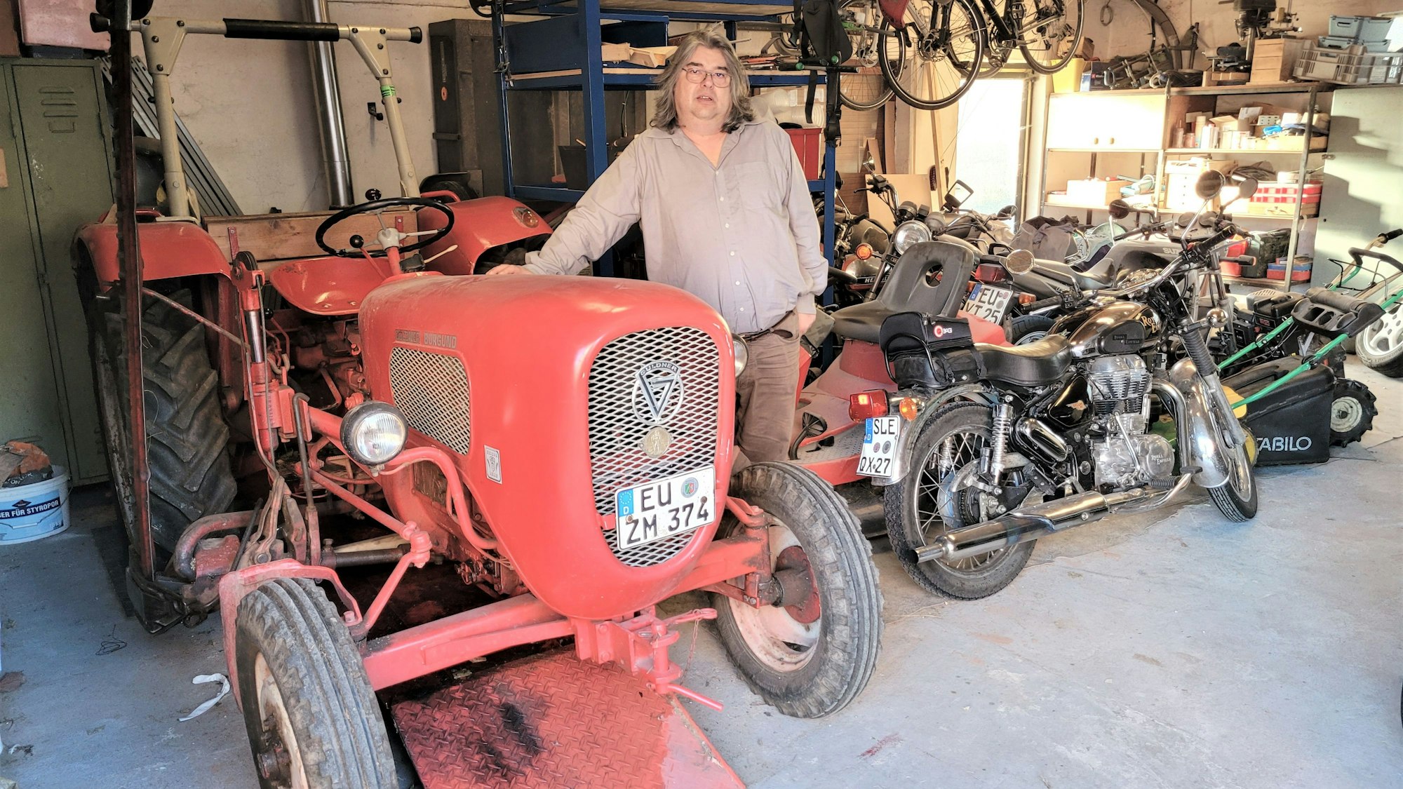 Ralf Meier aus Dottel steht in einer Garage neben einem roten Traktor und einem Motorrad.