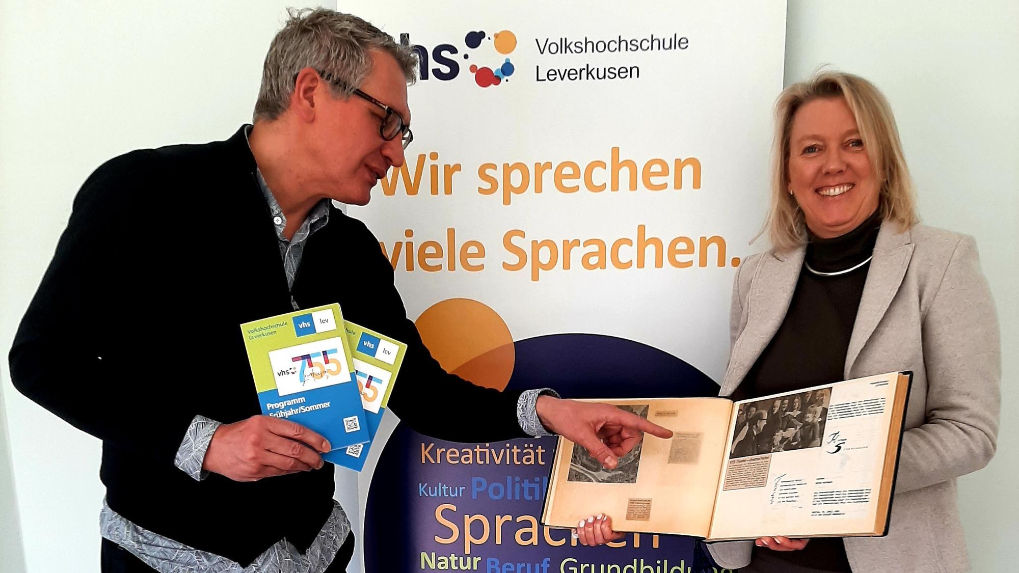 Günter Hinken, Leiter der Volkshochschule (VHS) Leverkusen, und seine Stellvertreterin Ute Wagner zeigen das VHS-Programm und ein Fotoalbum vor einem VHS-Plakat.