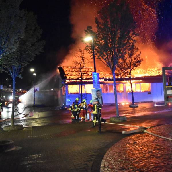 Eine Markthalle steht nachts lichterloh in Flammen. Feuerwehrleute versuchen mit Wasser, die Flammen zu löschen.