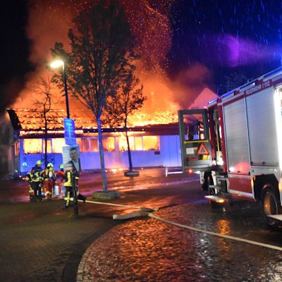 Eine Markthalle steht nachts lichterloh in Flammen. Feuerwehrleute versuchen mit Wasser, die Flammen zu löschen.