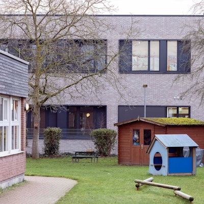 Es ist ein grauer verklinkerter Schulbau zu sehen. Davor ein Rasen mit Spielgeräten und einem blauen Schuppen.