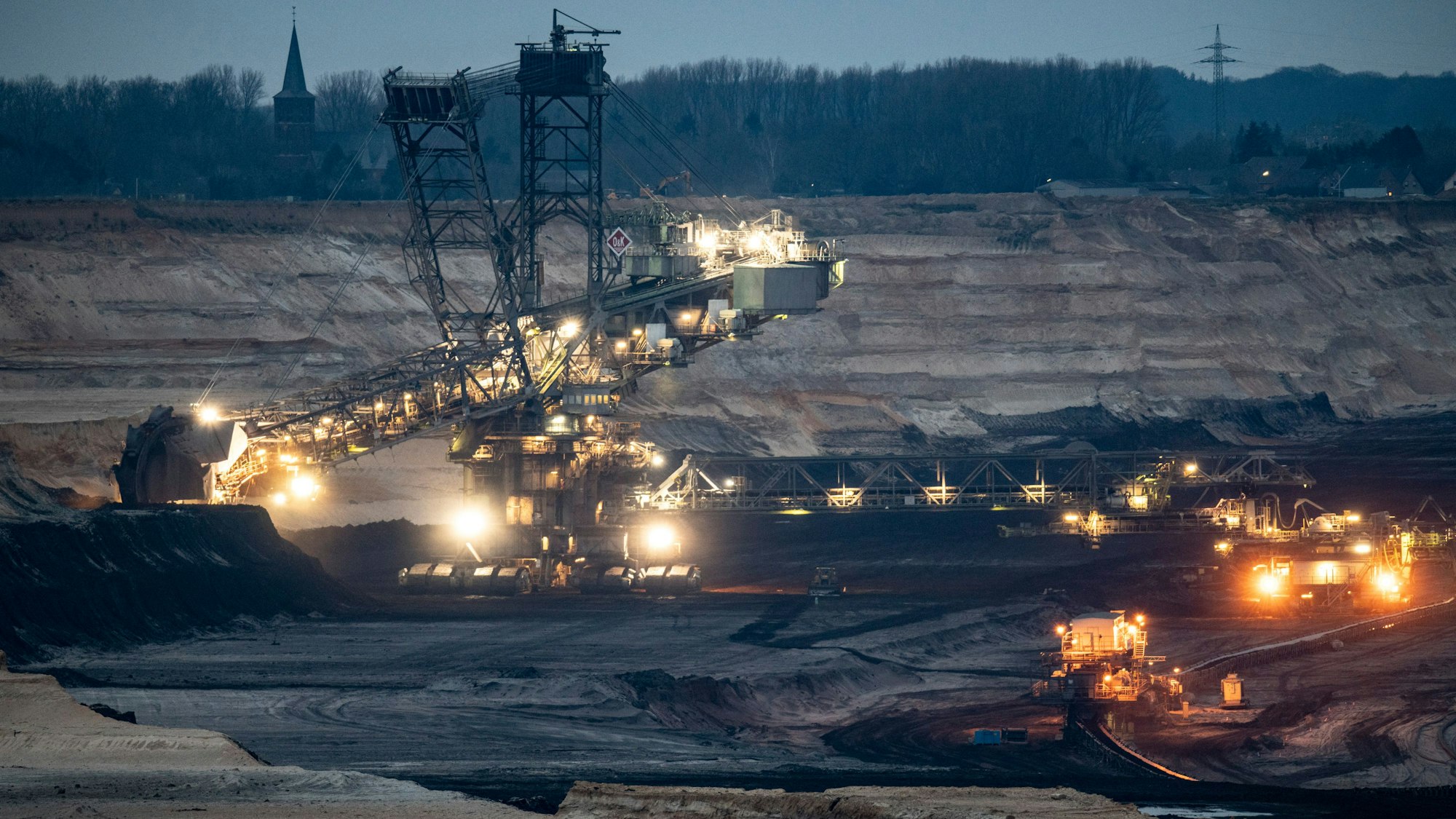 Ein riesiger Bagger im Tagebau Garzweiler. Im Hintergrund ist ein Kirchturm zu erkennen. Das Foto ist am Abend aufgenommen, zahlreiche Lichter leuchten.