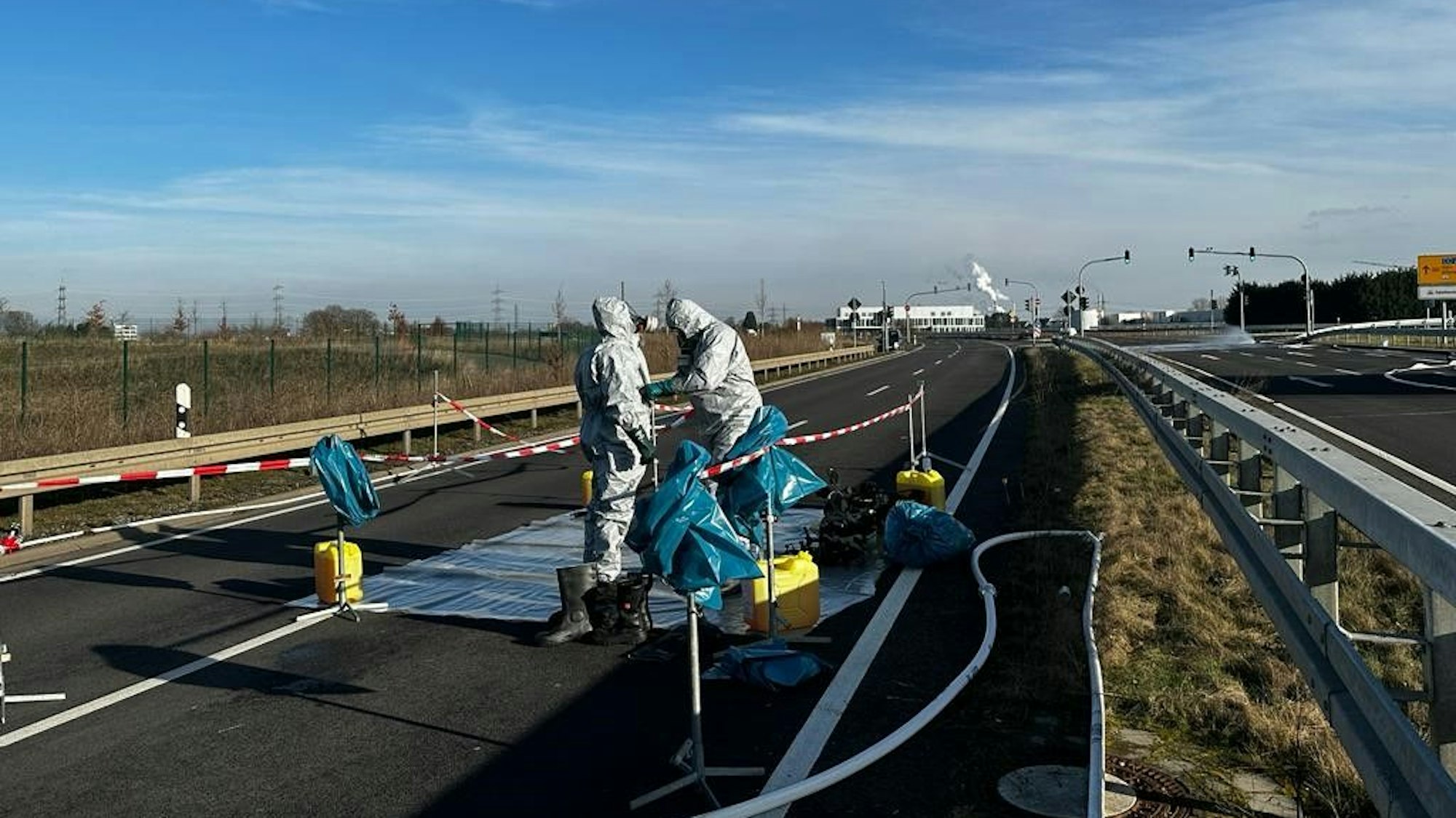 Feuerwehrleute kümmern sich um die ausgetreten Flüssigkeit auf der B 265n in Hürth. Sie tragen weiße Schutzanzüge.
