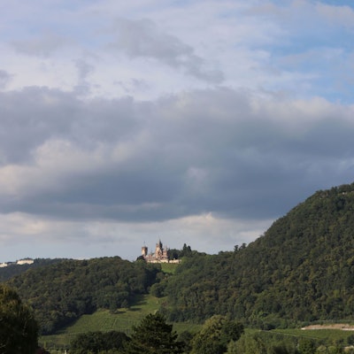 Das Bild zeigt eine kleine Hügelkette im Sommer mit grünen Bäumen und auf den Bergkuppen die Gebäude Petersberg-Hotel und Schloss Drachenburg sowie die Ruine des Drachenfels.