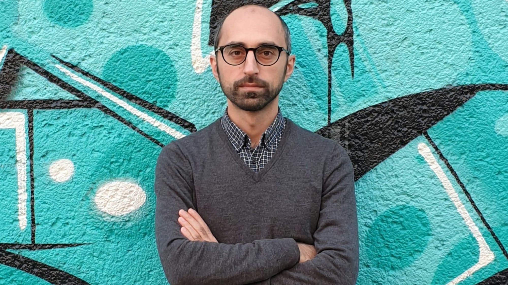 Eren Güvercin steht vor einer Mauer mit türkisem Graffiti. Er trägt eine Brille, ein kariertes Hemd und eine grauen Pullover.