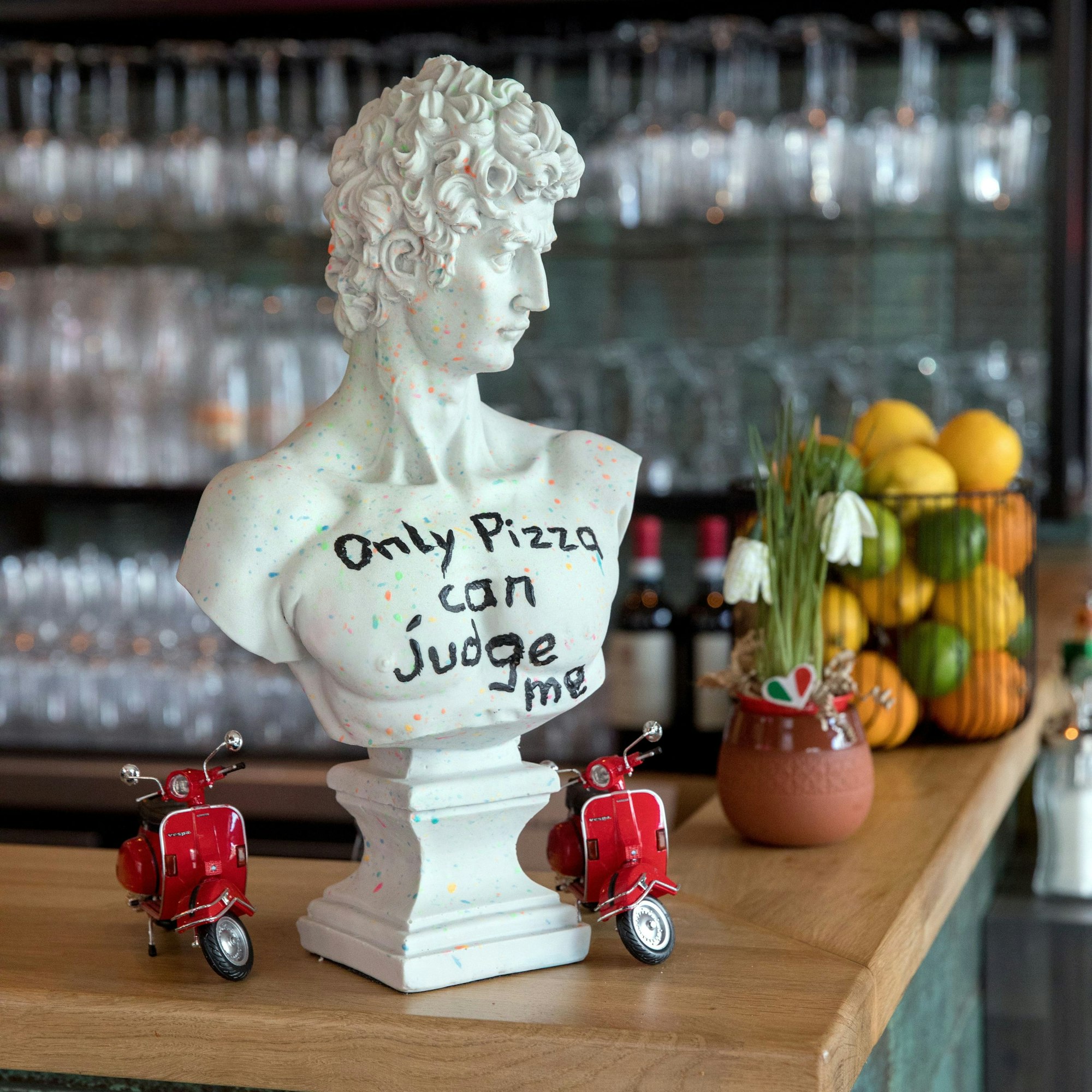 Ein kleine Statue mit der Aufschrift „Only Pizza can judge me“ - „Nur Pizza kann mich verurteilen“. Daneben zwei Modell-Vespas.