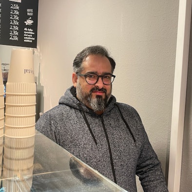 Der Besitzer von Back Löwe, Farshid Sowlati, steht hinter der Theke. Neben ihm stehen Mehrwegbecher der Kölner Firma Vytal. Seit dem 1.1.2023 müssen Gastronomen Mehrwegbehälter zum Mitnehmen anbieten, die nicht teuerer sein dürfen als Einwegverpackungen.